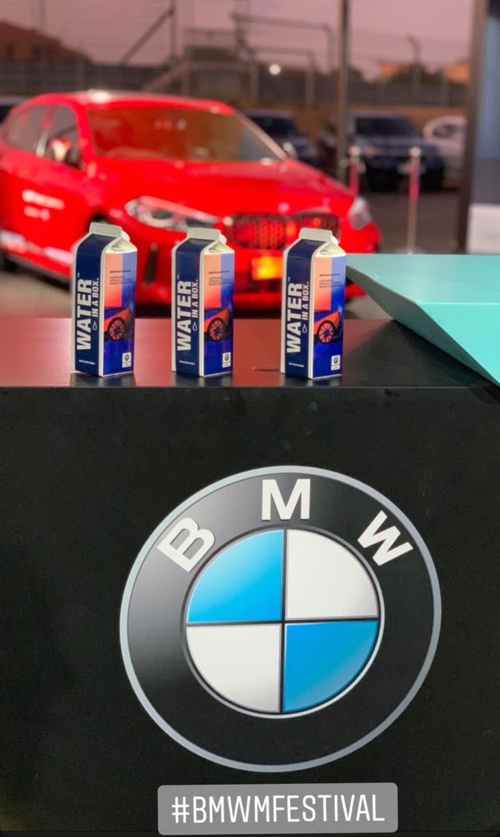 Three custom BMW M Festival Water In A Box cartons on a counter, with a red BMW behind