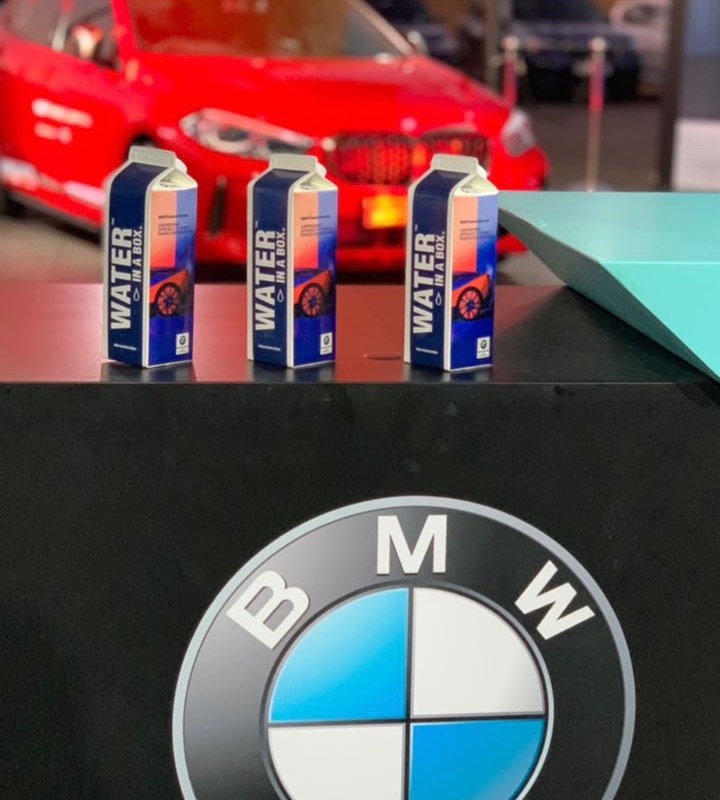 Three custom BMW M Festival Water In A Box cartons on a counter, with a red BMW behind