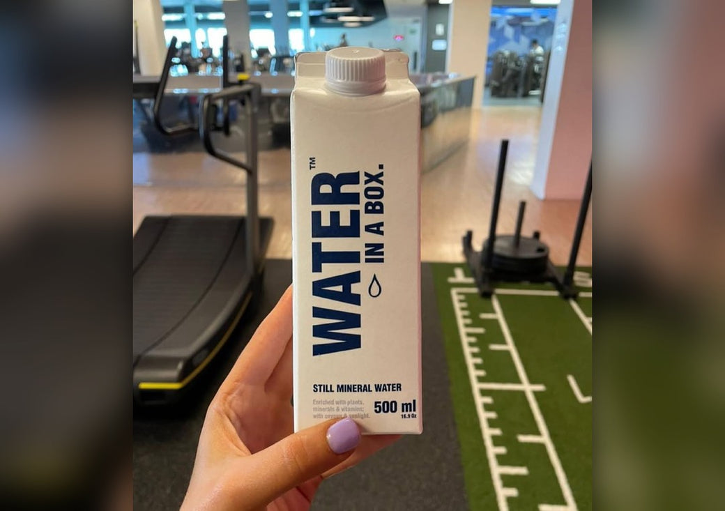 Hand holding a Water In A Box carton in a gym, with a treadmill and sled track behind